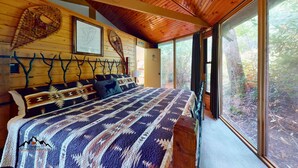 A bedroom with a large bed, a wooden headboard, and a view of the forest through large windows.