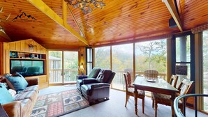A cozy cabin interior with a wooden ceiling, a television, a sofa, a dining table with chairs, and large windows offering a view of the outdoors.