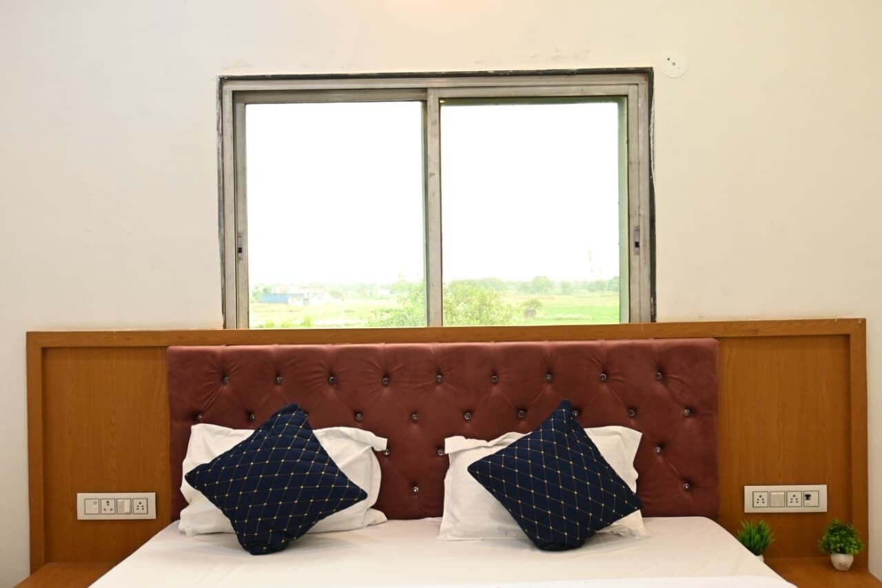 Une chambre à coucher avec une tête de lit en bois, deux oreillers blancs, deux oreillers bleu marine à motifs et une fenêtre donnant sur un paysage verdoyant.