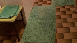 Un baño con bañera, lavamanos e inodoro. Hay alfombras verdes en el suelo y un adorno con forma de flor roja en la pared.