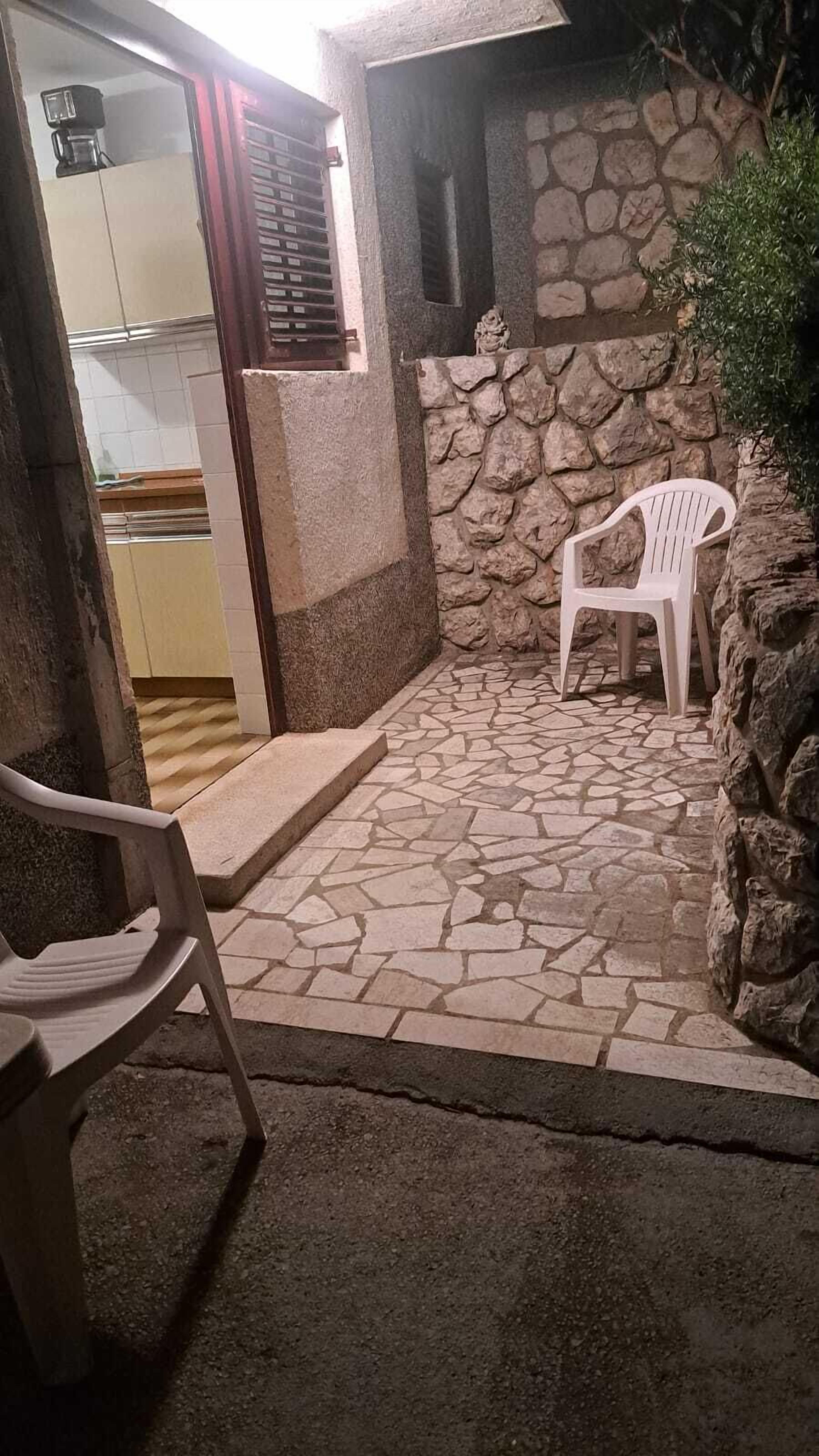 A stone-tiled patio with a white plastic chair, a small statue, and a kitchen area visible through an open door.
