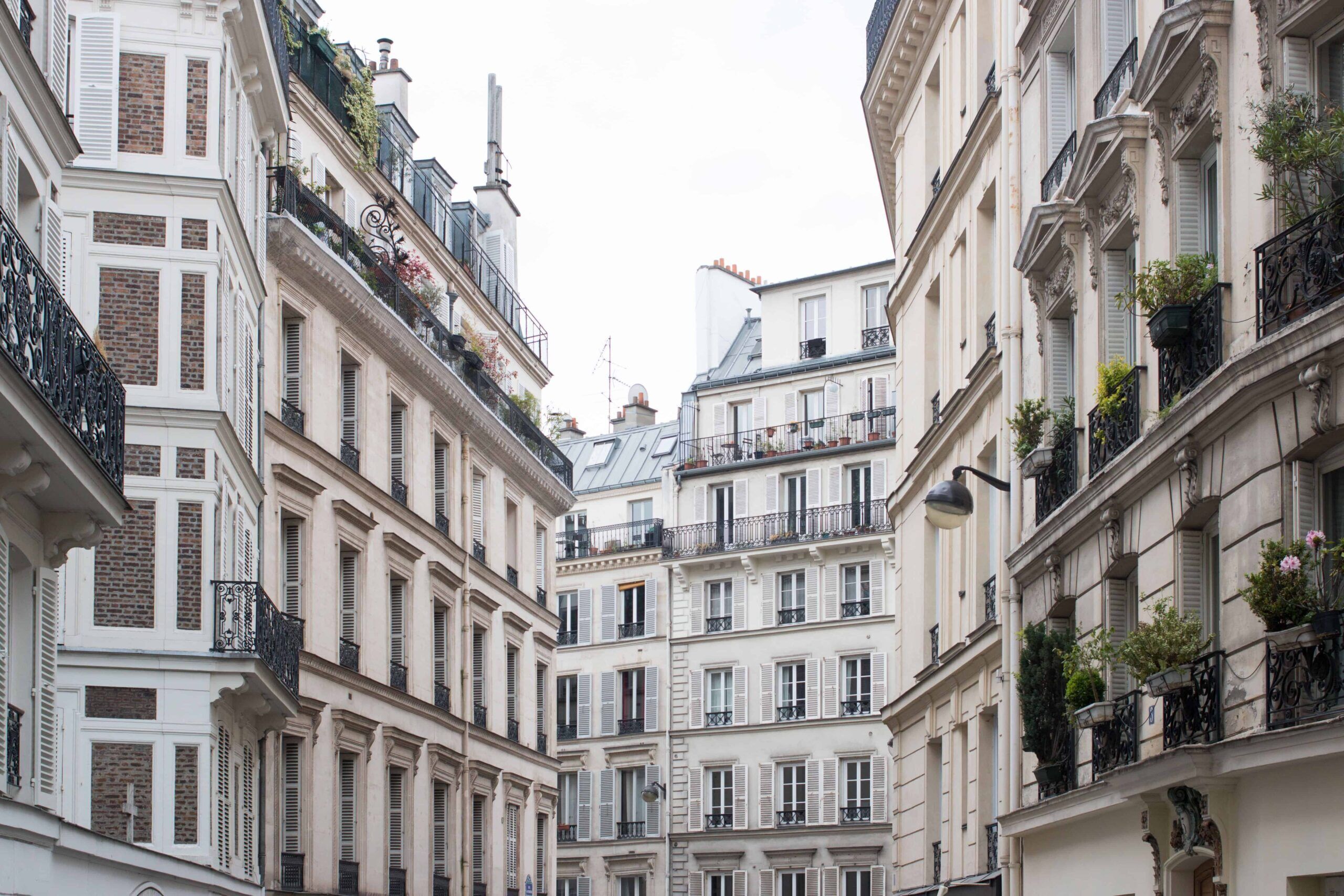 A narrow street with tall, closely packed buildings featuring balconies with railings and potted plants.