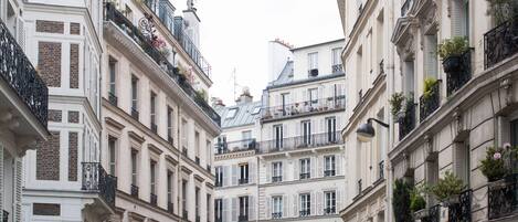 A narrow street with tall, closely packed buildings featuring balconies with railings and potted plants.