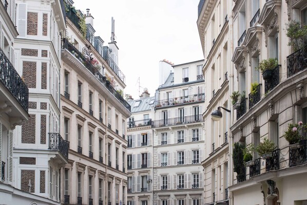 A narrow street with tall, closely packed buildings featuring balconies with railings and potted plants.