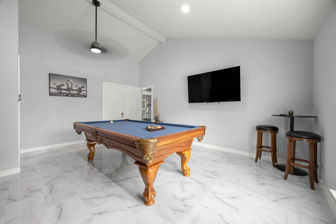 A pool table in a room with marble flooring, a ceiling light, a television on the wall, and two bar stools.