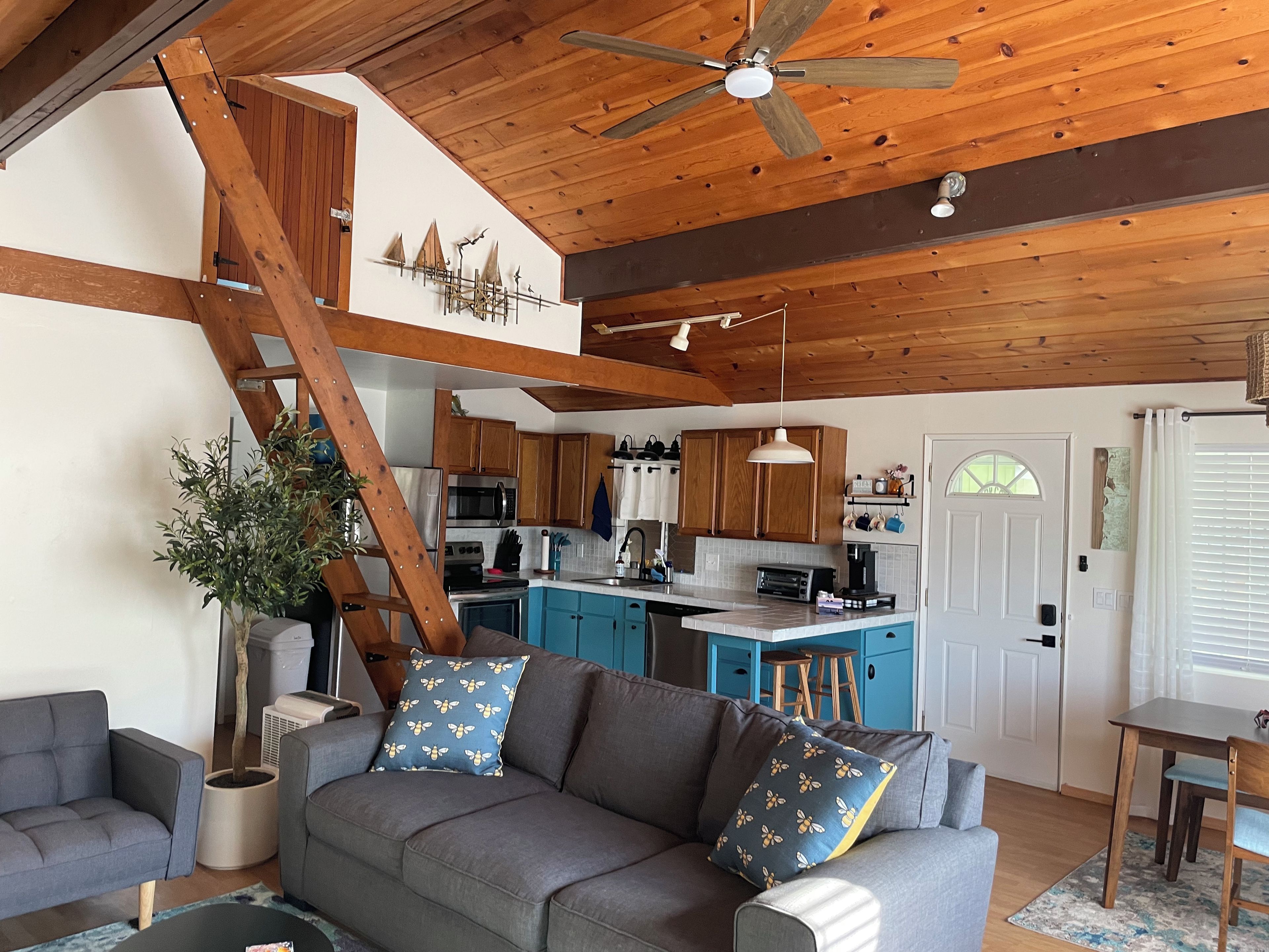 A cozy living room with a grey sofa, blue pillows, a wooden ceiling fan, and a kitchen area with wooden cabinets and a white door.