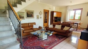 A living room with a staircase, a brown leather sofa, a coffee table with books, a red patterned rug, and a dining area with wooden furniture. - Spacious and cozy villa located between the sea and the volcano Etna! (Santa Maria La Stella)