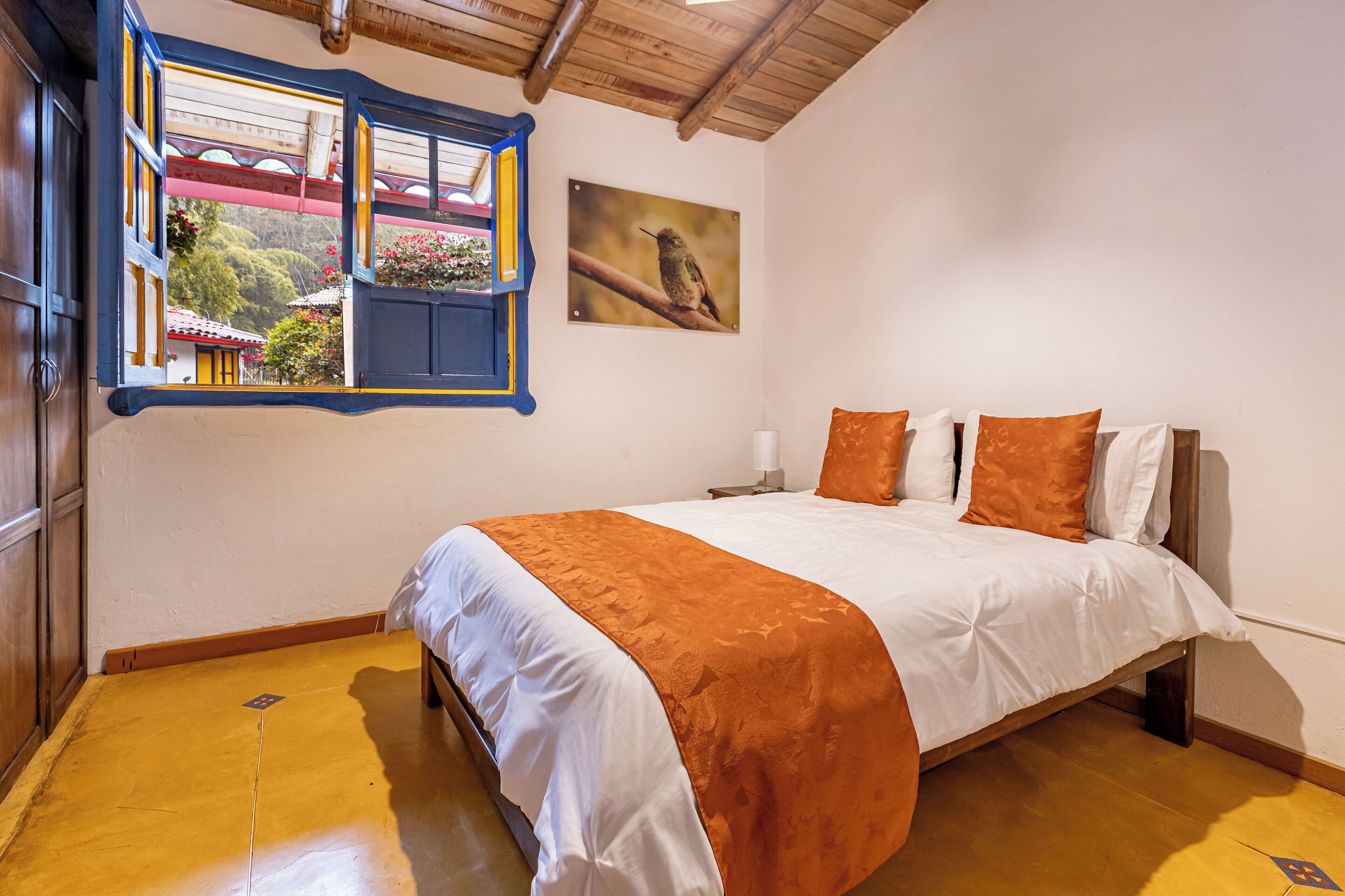 Een slaapkamer met een bed, een raam met uitzicht op een gebouw en bomen, en een schilderij van een vogel aan de muur.