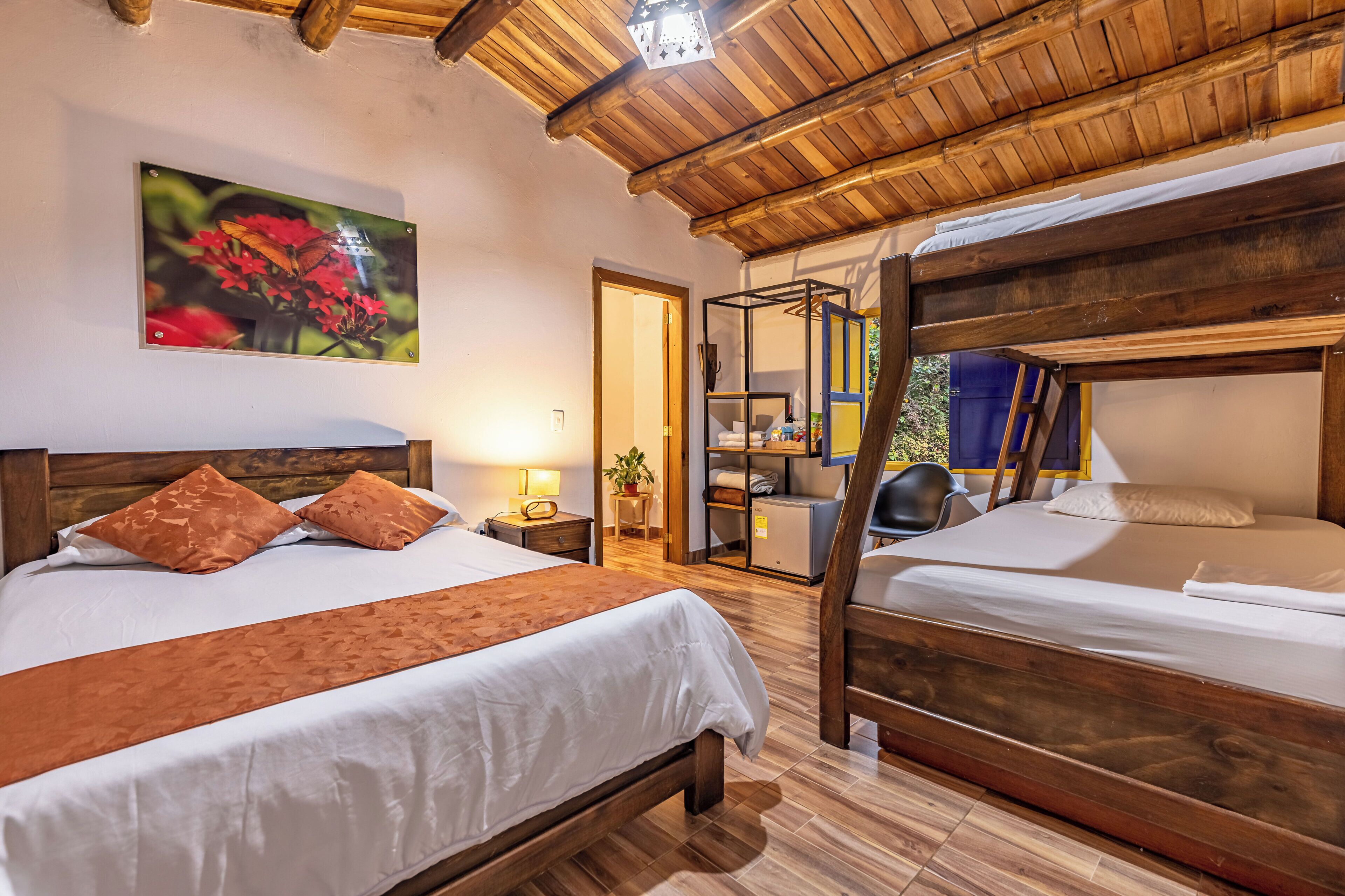 A room with a large bed, a smaller bed, wooden flooring, a wooden ceiling, a painting of flowers, a lamp, and a desk with a chair.