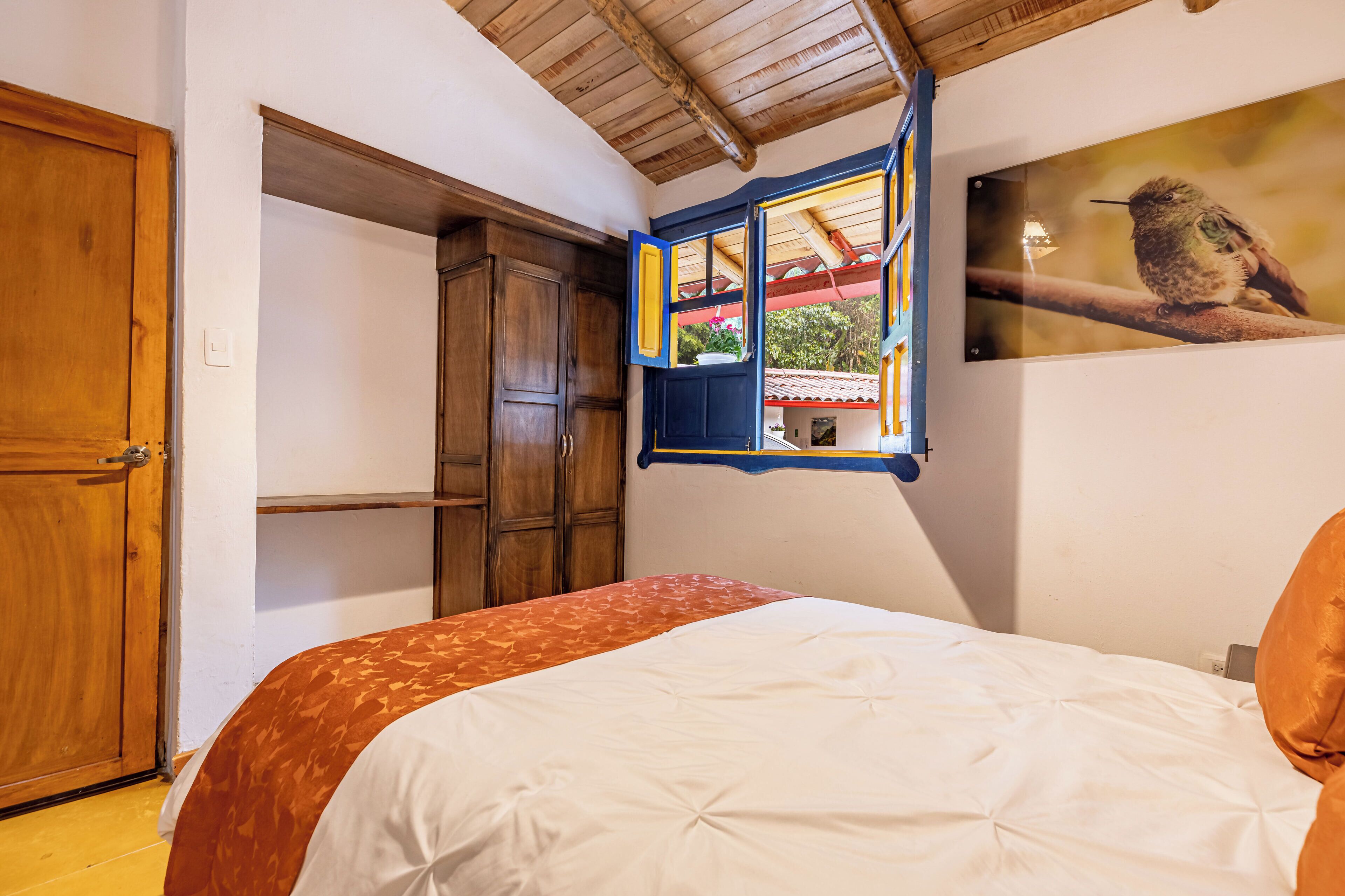 A bedroom with a bed, wooden wardrobe, and a window with a view of a house and greenery.