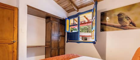 A bedroom with a bed, wooden wardrobe, and a window with a view of a house and greenery.