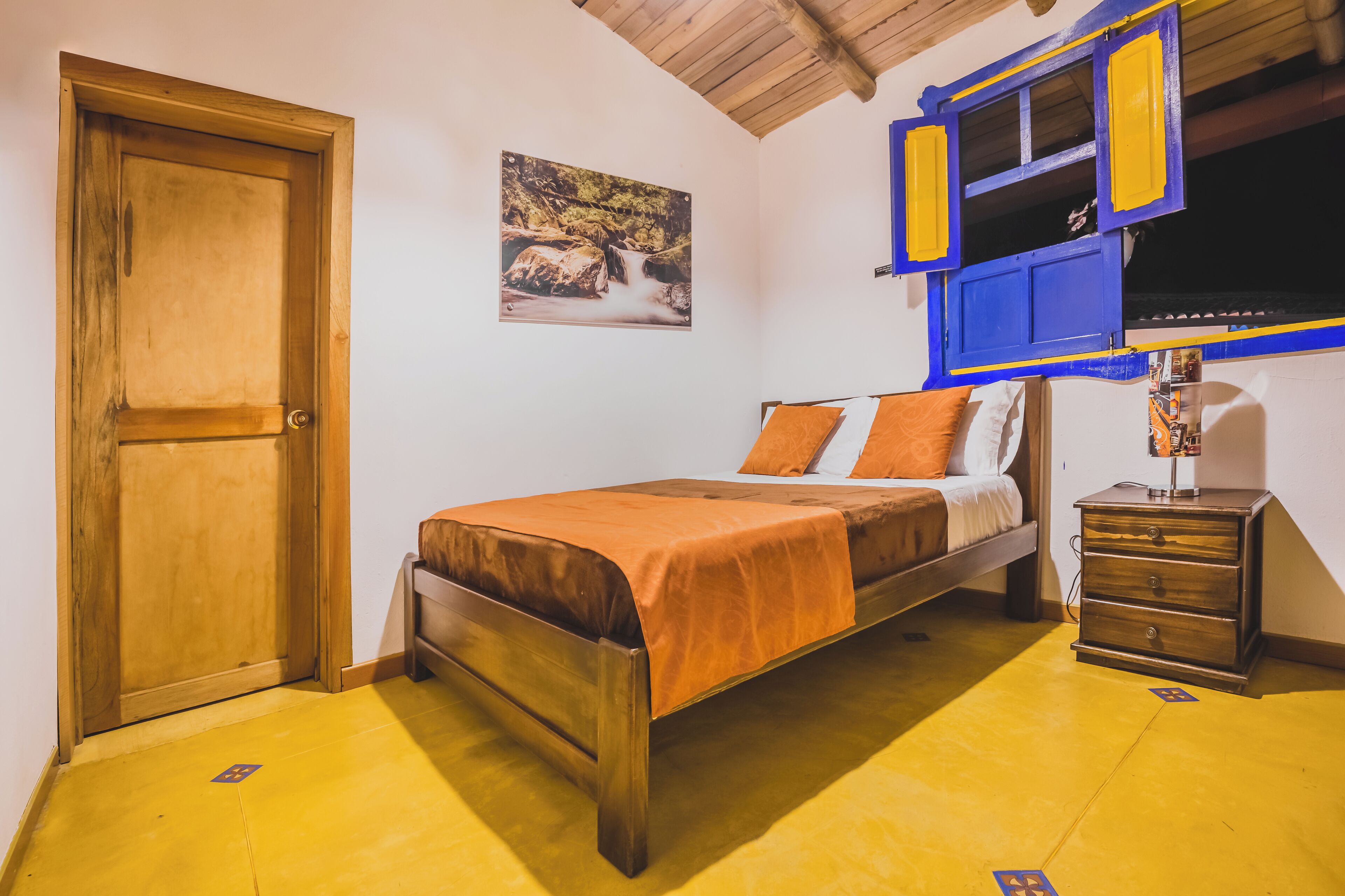 A bedroom with a wooden door, a bed with orange bedding, a nightstand, and a wall-mounted TV.