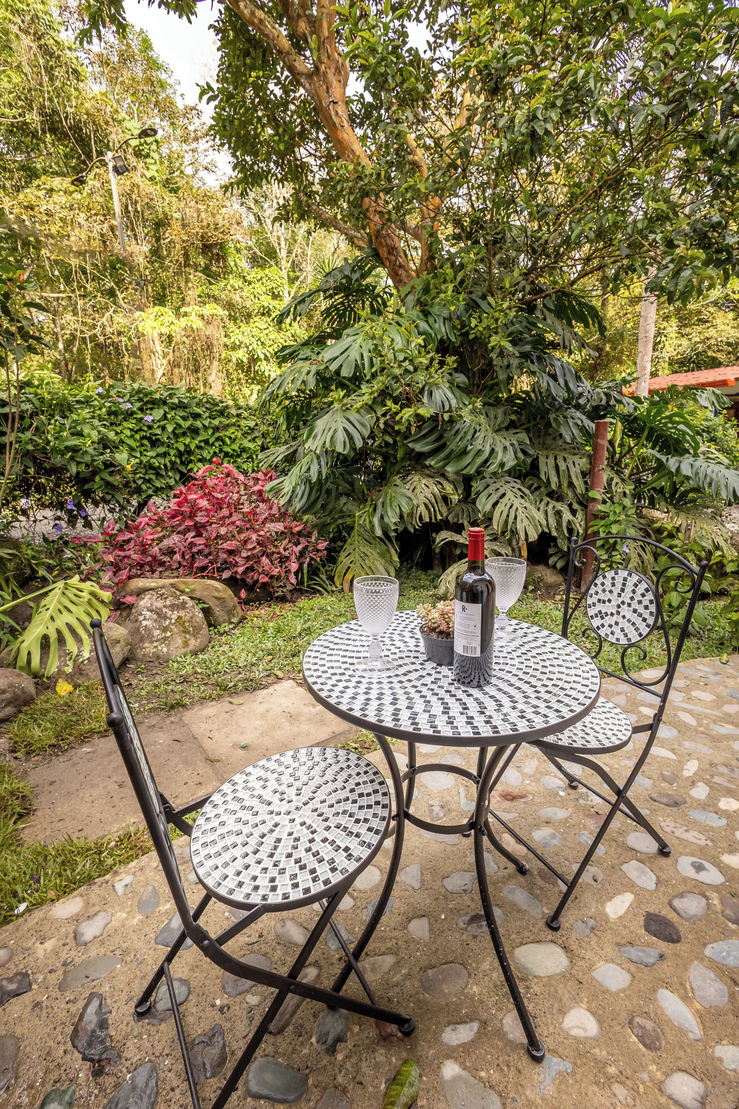 A round table with a wine bottle, surrounded by four chairs, set on a stone patio with a garden backdrop.