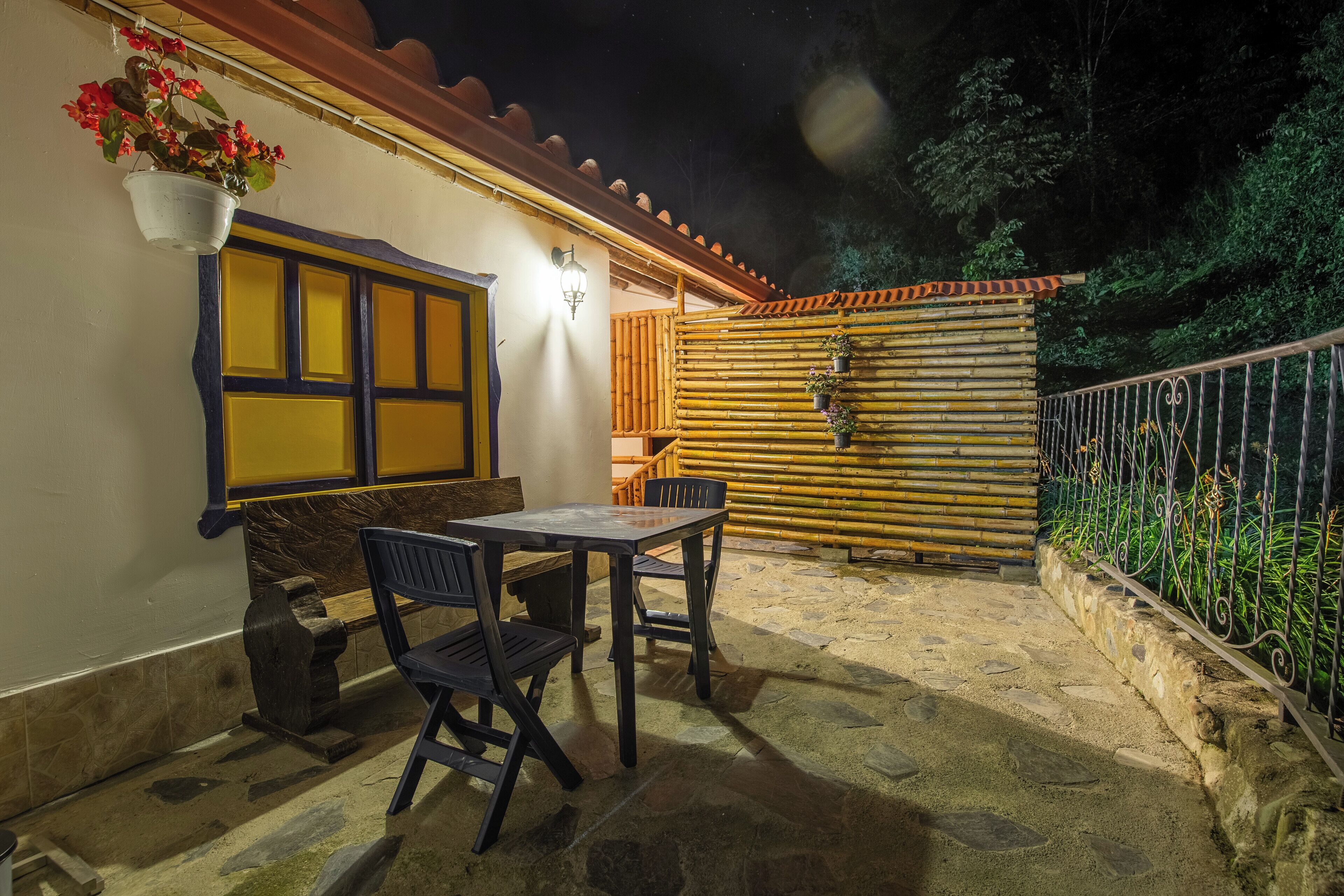 Une terrasse extérieure avec un sol en pierre, une table et des chaises en bois, un pot de fleurs suspendu et une clôture en bambou.