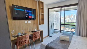 A hotel room with a bed, a desk with two chairs, a TV mounted on the wall, and a large window with a city view.