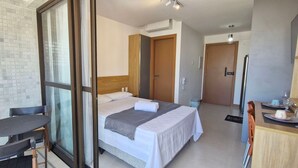 A hotel room with a bed, a small table with a chair, a glass door leading to a balcony, and a kitchenette area.