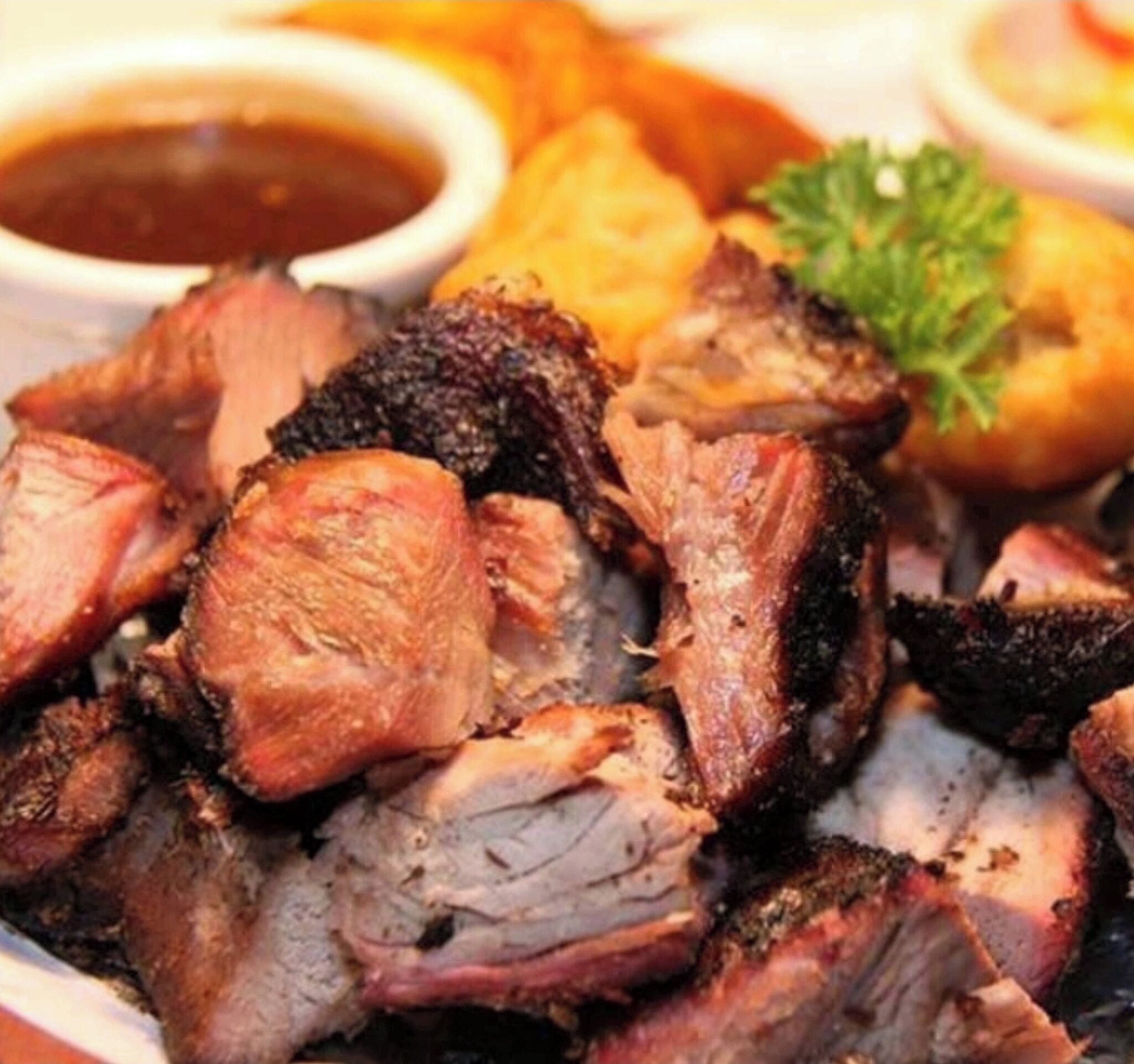 A close-up of sliced roasted meat, likely pork, with a charred exterior, served on a plate with a garnish of parsley.