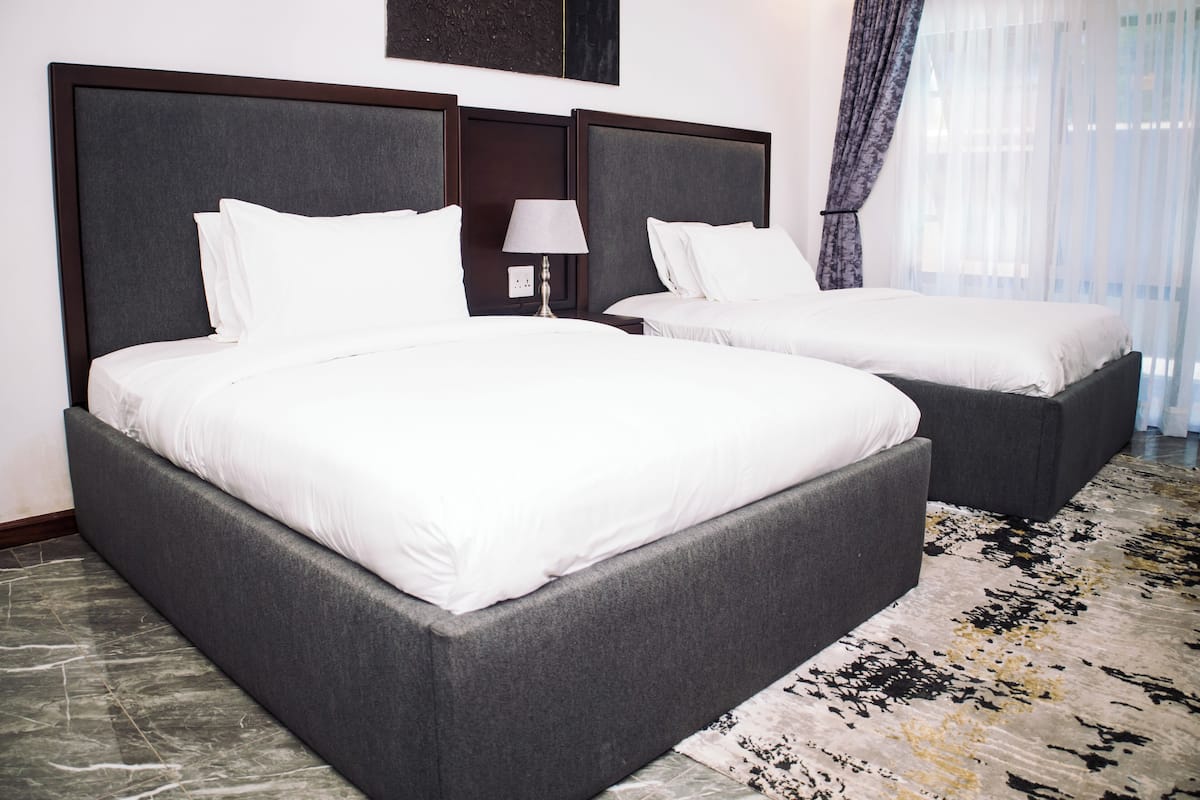A hotel room with two beds, a nightstand with a lamp, a marble floor, and a large window with curtains.