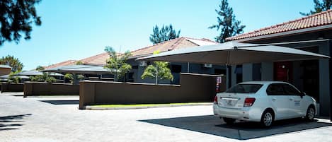 Un estacionamiento con un coche blanco, asientos al aire libre cubiertos y un edificio con tejado de tejas rojas.