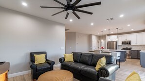 A modern living room with a black leather sofa, a brown ottoman, and a black armchair with yellow cushions. A ceiling fan and recessed lighting are visible.