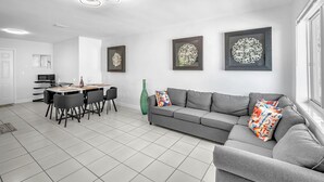 A modern living room with a grey sofa, a dining table with chairs, and a large window.
