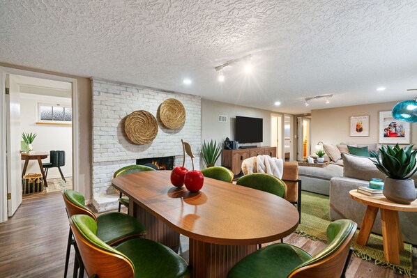 Una sala de estar moderna con una mesa de comedor de madera, sillas tapizadas en verde, una pared de ladrillo blanco y una chimenea.
