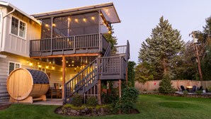 Una casa de dos pisos con un balcón, un barril de madera y un patio con luces colgantes.