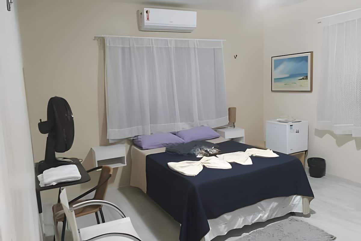 A room with a bed, a massage chair, a desk, a window with curtains, a wall-mounted air conditioner, and a picture on the wall.