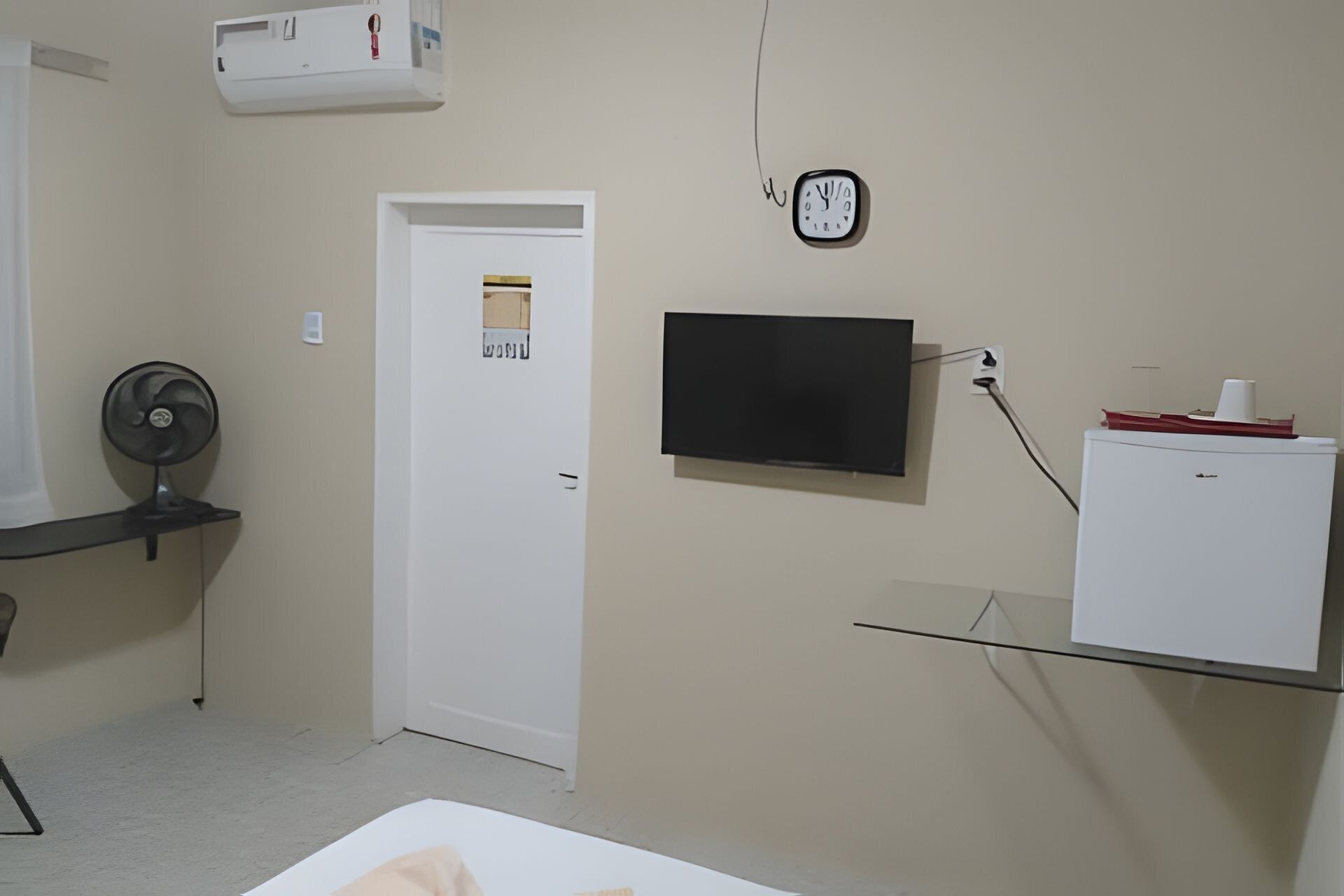 A room with a white door, a wall-mounted clock, a television, and a small refrigerator.