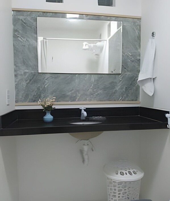 Un baño con lavamanos de mármol, un espejo, un florero con flores y un cubo de basura blanco.