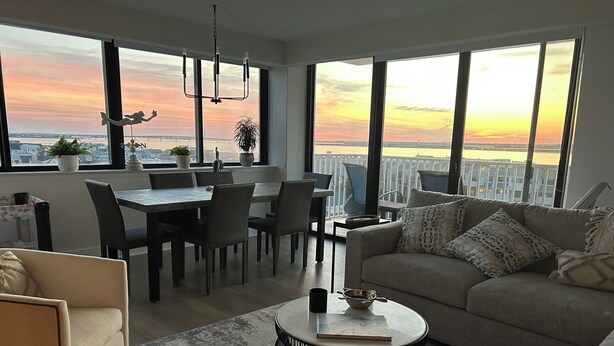 En moderne stue med spiseområde, store vinduer med udsigt til solnedgang og en balkon.