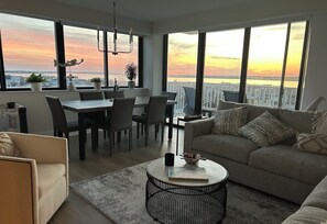 A modern living room with a dining area, large windows with a sunset view, and a balcony. - Luxury LONGPORT Condo Amazing Views (Longport)