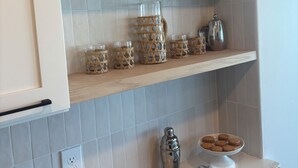 Et køkken med hvide skabe, en marmor bordplade og et flisebeklædt bagpanel. Der er hylder med dekorative genstande, en kaffemaskine og en tallerken med småkager.
