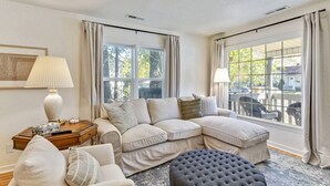 A living room with a sectional sofa, a round ottoman, a side table with a lamp, and large windows with curtains. - Nest & Nook • 2 Units, 2 Mins from BYU (Provo)