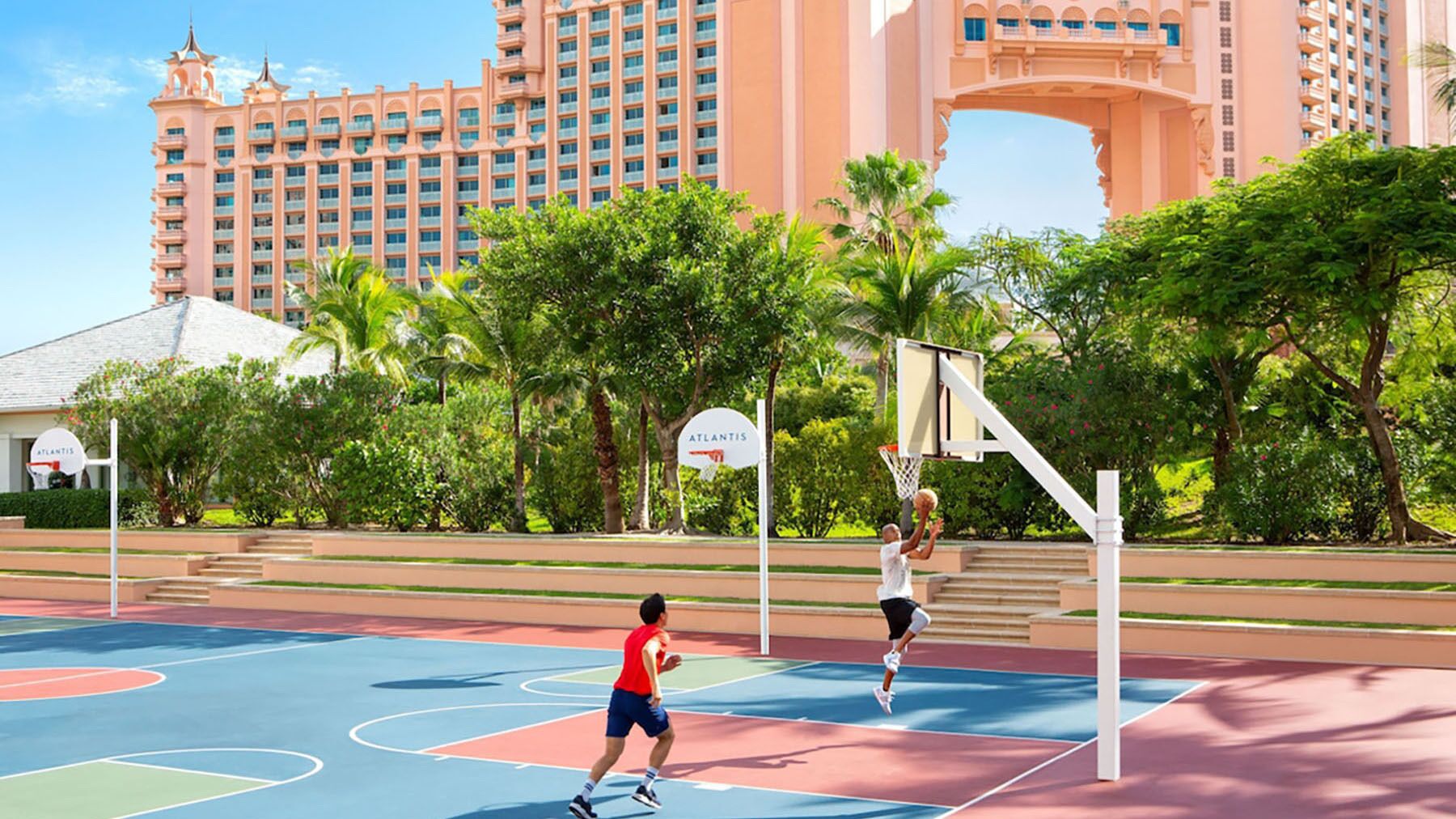 A basketball court with two players, surrounded by lush greenery and a large hotel building in the background.
