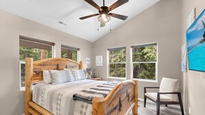 Un dormitorio con cama de madera, ventanas amplias, ventilador de techo y una silla.