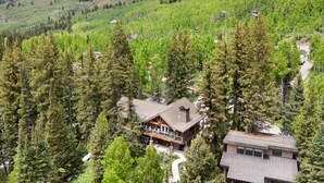Una cabaña con una amplia terraza y un edificio separado, rodeada de un bosque denso y colinas.