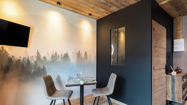 A modern dining area with a table set for two, a mounted TV, and a wall mural of a forest. - Cosy studio in downtown Gérardmer (Gérardmer)