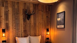 A bedroom with a wooden wall, a mounted deer head, a bed with white bedding, and two wall-mounted lamps. - Cosy studio in downtown Gérardmer (Gérardmer)