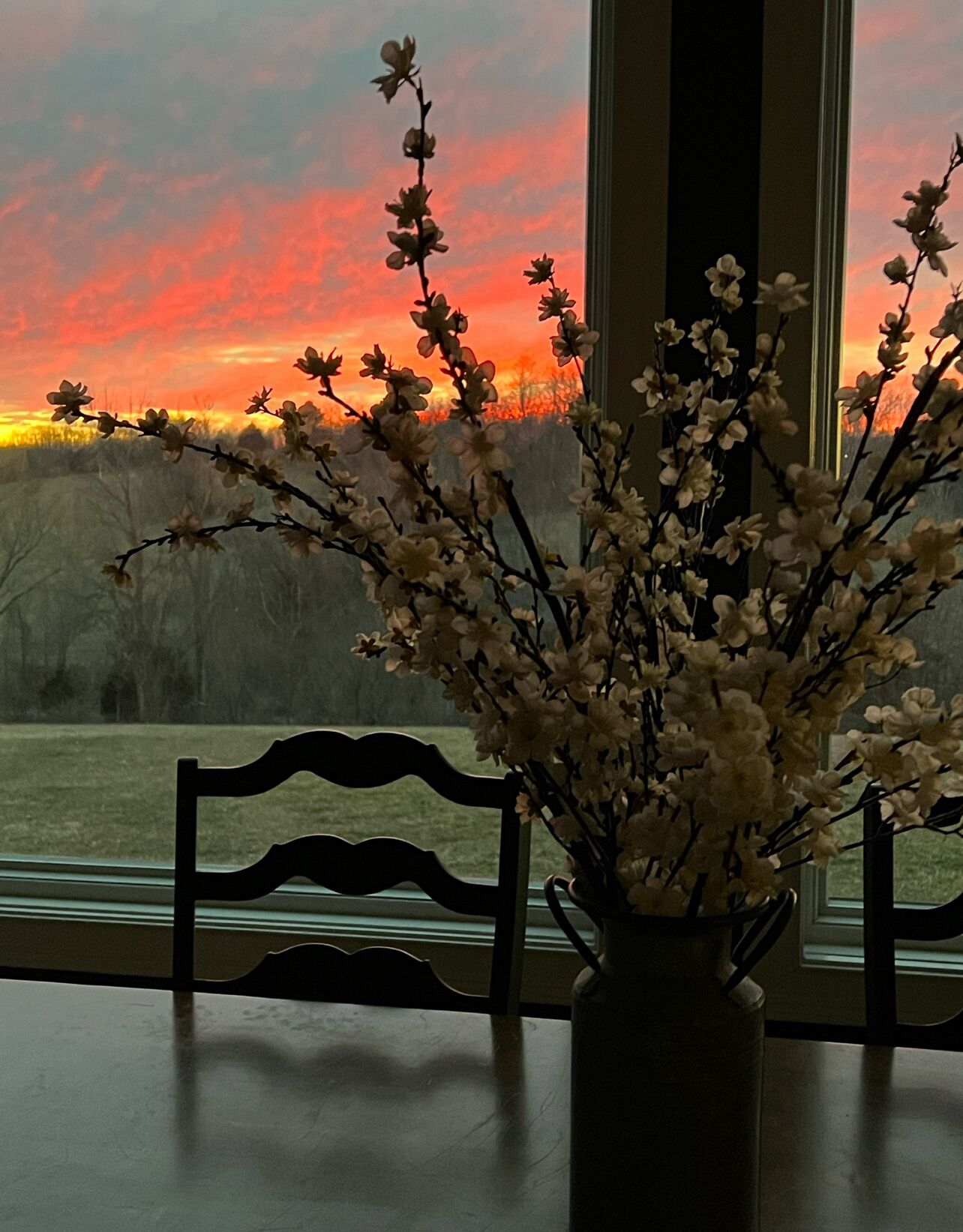 Um vaso com flores sobre uma mesa perto de uma janela com vista para o pôr do sol.