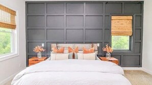 Quarto com cama grande, cabeceira escura em painéis, duas mesas de cabeceira com luminárias e janela com vista para o verde.