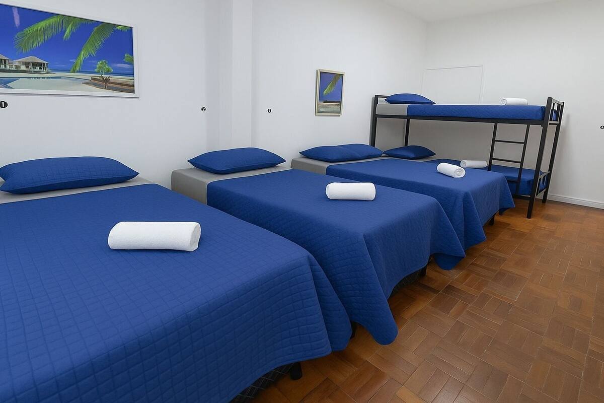 A hotel room with four single beds, each with a blue bedspread and a white towel. There is a bunk bed in the corner with a blue mattress. The room has white walls, a wooden floor, and a painting of a beach scene on the wall.