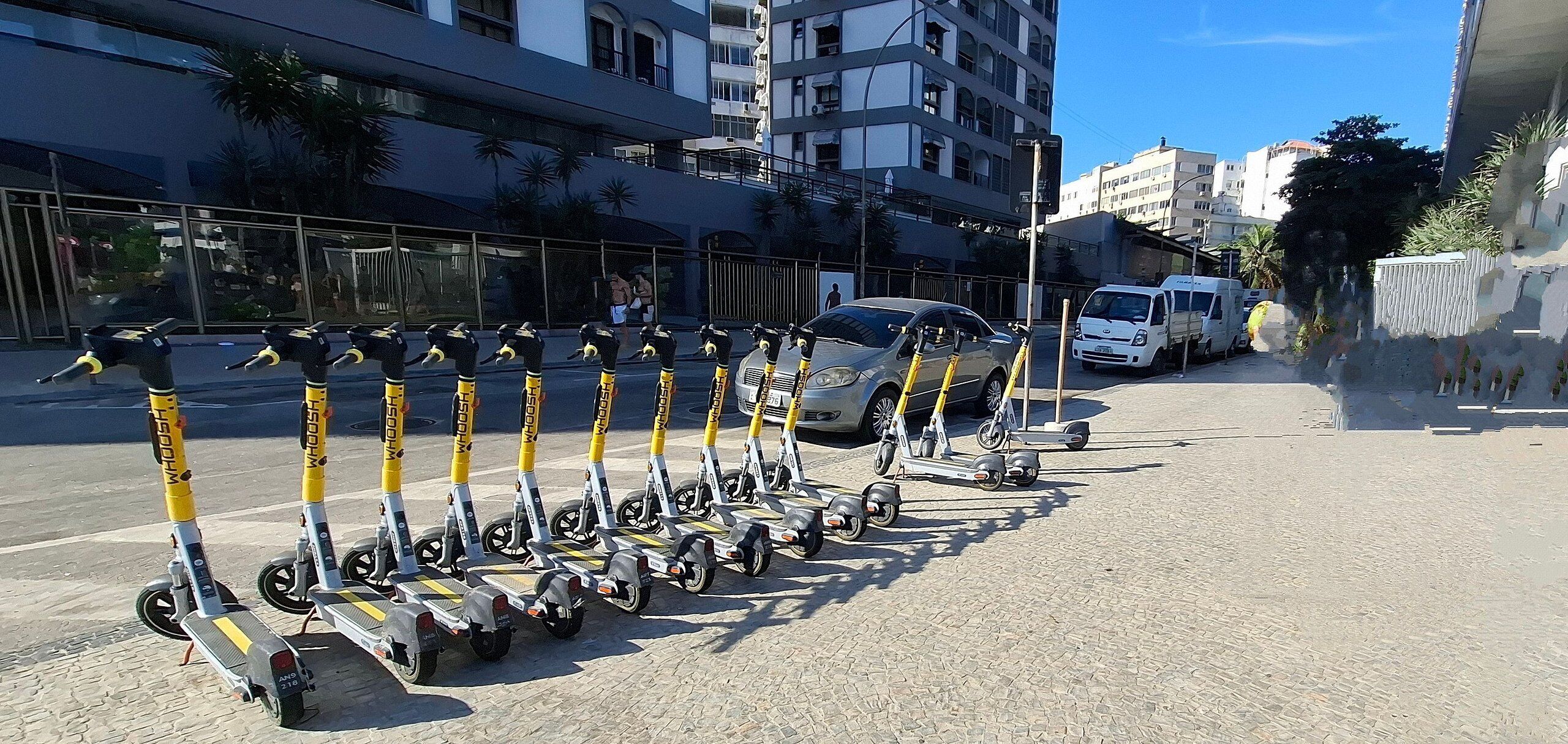A row of electric scooters parked on a paved area in front of a building with multiple floors and balconies.
