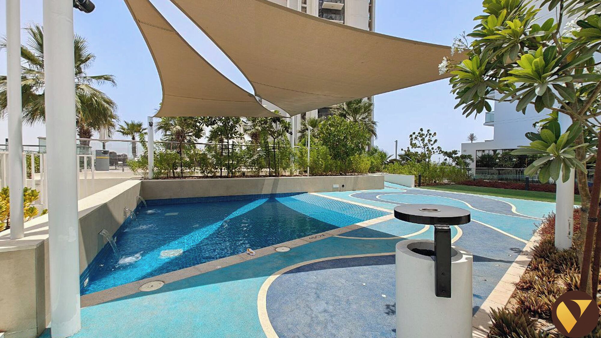 A swimming pool with a blue and white tiled design, surrounded by palm trees and a shaded area.