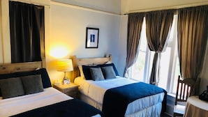 A hotel room with two beds, a bedside table with a lamp, a framed picture on the wall, and a window with curtains. - Hotel San Ramon (Bisbee)