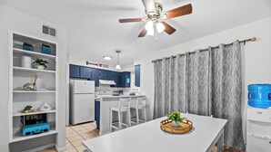 Une cuisine moderne avec des meubles blancs, une table à manger avec des chaises, un ventilateur de plafond et un distributeur d’eau.