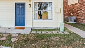 Un immeuble de deux étages avec une porte bleue, un petit jardin devant et un mur de briques sur le côté droit.