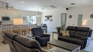 Ein modernes Wohnzimmer mit einer Couch, einem Sessel und einem Couchtisch. Es gibt eine Küchenzeile mit einer Mikrowelle und Stühlen.