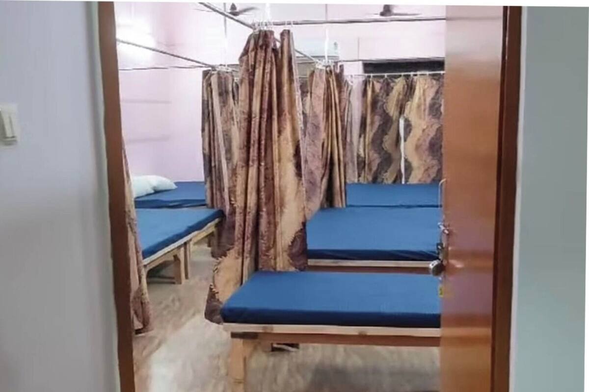 Ein Zimmer mit vier Betten, jedes mit blauer Matratze und Holzrahmen. Vorhänge hängen von der Decke und ein weißes Kopfkissen ist auf einem Bett sichtbar.