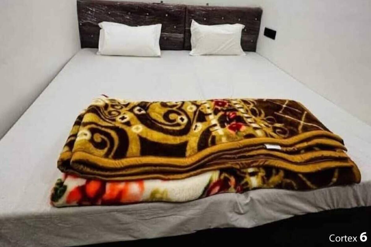 Ein Bett mit einer gemusterten Decke, zwei weißen Kissen und einem dunklen Kopfteil in einem Raum mit schlichten Wänden.